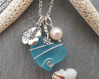 Puede incluir: Un collar de cadena de plata con un colgante de corazón de vidrio azul envuelto en alambre de plata. El colgante también presenta un amuleto de hibisco plateado y una perla blanca.
