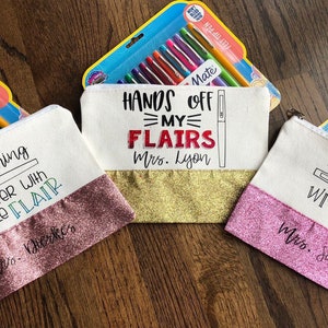 May include: Three white canvas zippered pouches with pink, gold, and pink glitter accents. The pouches have different sayings on them: "Everything is better with a little flair", "Hands off my flairs", and "Teaching with flair".