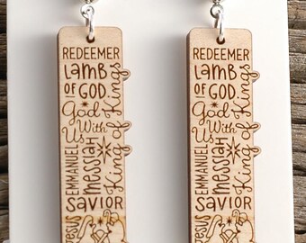 May include: A pair of wooden earrings with a silver ball stud. Each earring is a light-colored rectangle with engraved text including "Redeemer, Lamb of God, Jesus, Savior." The earrings are displayed on a white card.