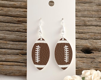 May include: A pair of football-shaped earrings. The earrings are brown with white stitching and accents, and are attached to silver-colored hooks. The earrings are displayed on a white card against a wooden background.