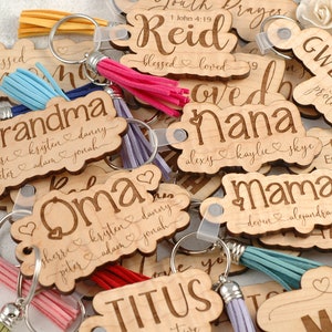 May include: A collection of wooden keychains with various names and phrases engraved on them. Each keychain has a small tassel attached. The keychains are arranged in a pile, with some of the tassels hanging over the edges.