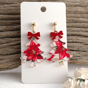 May include: Christmas tree-shaped earrings with a poinsettia design. The earrings feature a red bow at the top and gold-colored hardware. The poinsettia design is red and white, with green leaves.