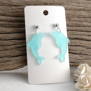 May include: A pair of light blue dolphin-shaped earrings. Each earring features a silver-colored ball stud and a translucent dolphin pendant. The earrings are displayed on a white card with a hole at the top, against a wooden background.