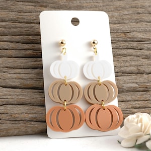 May include: Pumpkin-shaped earrings in white, tan, and orange, hanging from gold-colored posts. The earrings are displayed on a white card against a wooden background. The earrings are a fall-themed accessory.