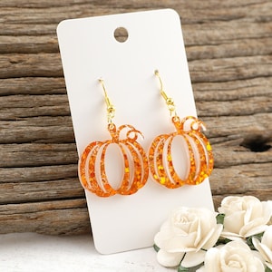 May include: A pair of orange glitter pumpkin earrings with gold-colored hooks, displayed on a white card. The earrings have a festive design, perfect for autumn or Halloween. The background features a wooden surface and white roses.