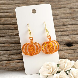 May include: A pair of orange glitter pumpkin earrings with gold-colored hooks. The earrings are displayed on a white card, with a wooden background. The earrings are a festive accessory.