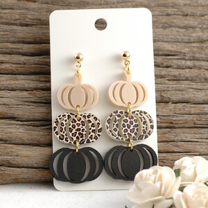 May include: A pair of dangle earrings with a fall theme. Each earring features three pumpkin shapes: a beige top, a leopard print middle, and a black bottom. The earrings are attached to a gold-colored post.