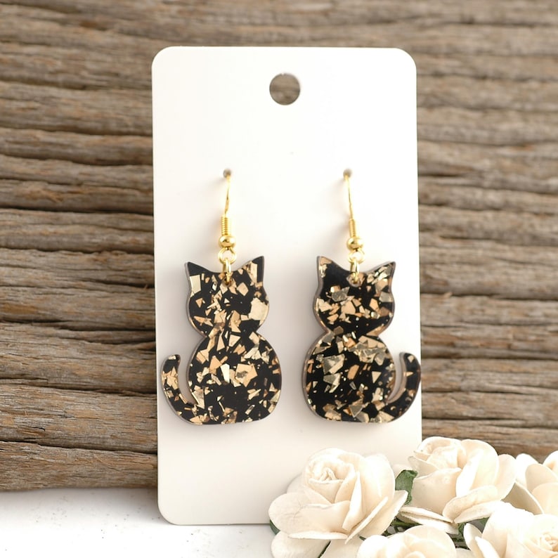 May include: A pair of black cat-shaped earrings with gold flecks, hanging from gold-colored hooks. The earrings are displayed on a white card against a wooden background, with white flowers in the foreground. The earrings are a fun accessory.
