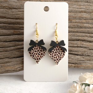 May include: A pair of heart-shaped earrings with a leopard print design. Each earring features a black bow at the top and gold-colored hooks. The earrings are displayed on a white card against a wooden background.