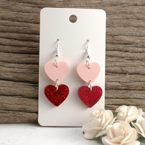 May include: A pair of heart-shaped earrings. Each earring features a light pink heart connected to a red glitter heart. The earrings hang from silver-colored hooks and are displayed on a white card.