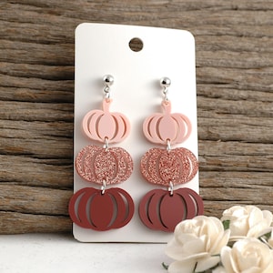 May include: Dangling earrings featuring a three-tiered pumpkin design. The top pumpkin is light pink, the middle is glittery rose gold, and the bottom is a deep reddish-brown. The earrings hang from silver-colored posts.
