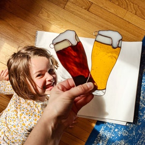 May include: Two stained glass beer glasses, one amber and one yellow, with white foam tops. The glasses are held up against a white background. The image is taken from a low angle.