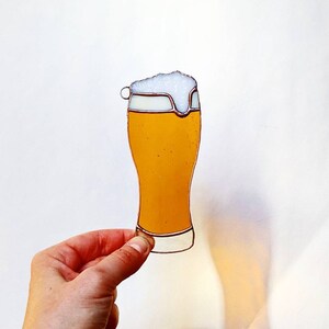 May include: A stained glass beer glass with a foamy head. The glass is filled with a golden-yellow liquid and has a clear border. The glass is held by a hand against a white background.