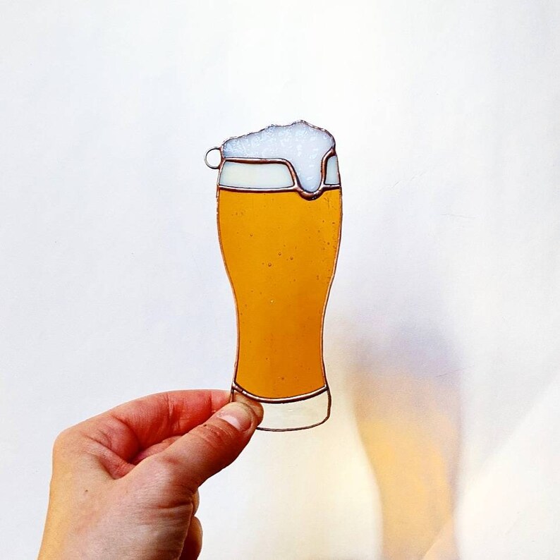 May include: A stained glass beer glass with a foamy head. The glass is filled with a golden-yellow liquid and has a clear border. The glass is held by a hand against a white background.