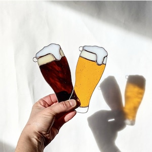 May include: Two stained glass beer glasses, one dark brown and one golden yellow, with white foam tops. The glasses are held against a white background, casting shadows. Each glass has a small metal loop at the top.