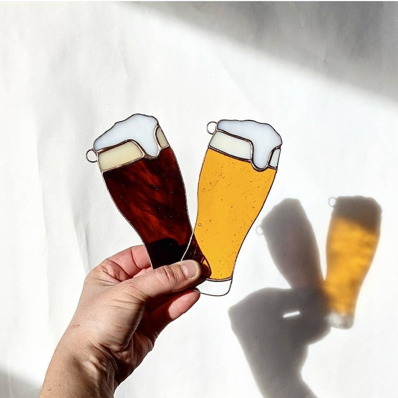 May include: Two stained glass beer glasses, one dark brown and one golden yellow, with white foam tops. The glasses are held against a white background, casting shadows. Each glass has a small metal loop at the top.