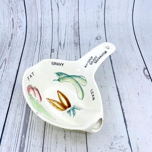 May include: A white ceramic gravy separator with a handle and a pouring spout. The separator has illustrations of vegetables and text that reads "FAT", "GRAVY", "LEAN", and "NU-DRIP GRAVY SEPARATOR".