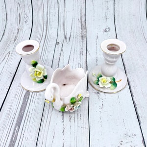 May include: A set of three white porcelain candle holders featuring a swan design with floral accents. The swan is a decorative dish with a floral design. The candle holders have a gold trim.