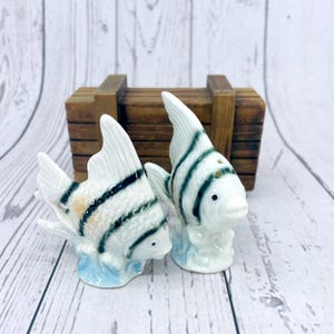 May include: A pair of white ceramic salt and pepper shakers shaped like fish with black and green stripes. The fish are sitting on a blue and white base.