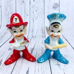 May include: Two vintage porcelain figurines of elves. One elf is dressed as a firefighter with a red hat and red trousers. The other elf is dressed as a police officer with a blue hat and blue trousers. Both elves are holding a small object in their hands.