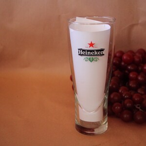 May include: A clear glass beer glass with a tapered shape, featuring the Heineken logo with a red star and black text on a white paper insert placed inside the glass. The glass is positioned next to a cluster of dark red grapes.
