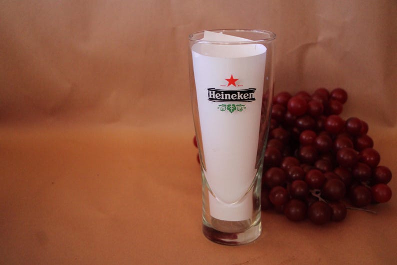 May include: A clear glass beer glass with a tapered shape, featuring the Heineken logo with a red star and black text on a white paper insert placed inside the glass. The glass is positioned next to a cluster of dark red grapes.