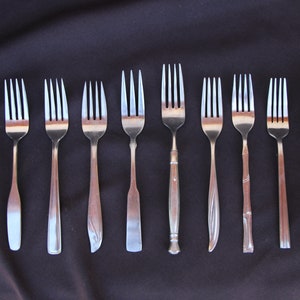 May include: Eight silver forks with different designs and styles laid out on a black surface.