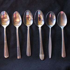 May include: Eight silver spoons with varying designs and finishes. The spoons are arranged in a row on a dark surface.