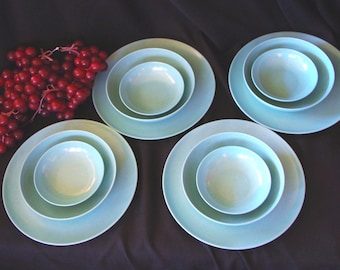 May include: A set of four aqua-colored ceramic dish sets, each with a large plate, a medium plate, and a small bowl. A cluster of red grapes is in the upper left corner. The dishes are arranged on a dark surface.