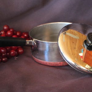 May include: A stainless steel saucepan with a copper-colored base and a black handle. The pan includes a matching stainless steel lid with a black knob. The exterior of the lid is highly reflective, showing a distorted reflection of the surroundings.