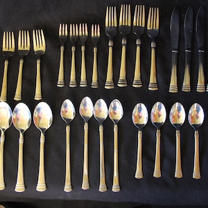 May include: A collection of vintage silverware, including forks, knives, and spoons, arranged on a dark surface. The silverware features gold-toned handles and silver-toned heads. The set includes various sizes of forks, knives, and spoons.