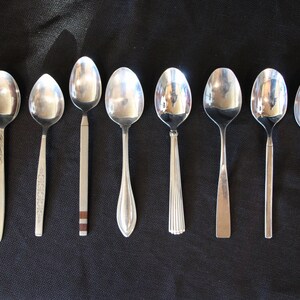 May include: A set of eight silver spoons with varying designs. The spoons are arranged in a row on a black surface.