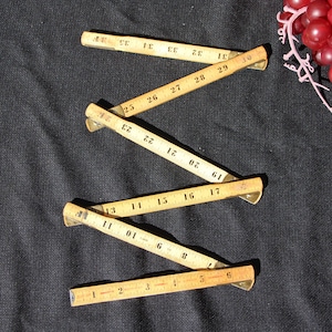 May include: A vintage wooden folding ruler, with black numbers and markings, is laid out on a dark background. The ruler is partially extended, showing measurements in inches. A cluster of red grapes is in the upper right corner.