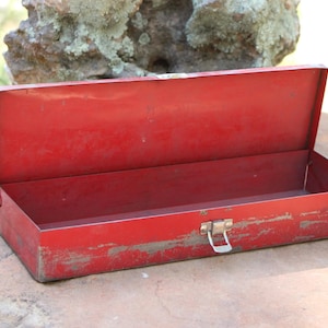 May include: A rectangular red metal tool box with a hinged lid and a silver-colored metal latch on the front. The exterior paint shows signs of wear and weathering, revealing patches of the underlying metal surface along the bottom edges.