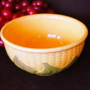 May include: A yellow ceramic bowl with a corn cob design. The bowl has a textured exterior and green leaves at the base. The interior is a lighter yellow. A bunch of red grapes is in the background.