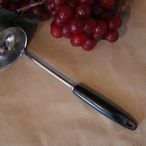 May include: A silver ladle with a black handle. The ladle is on a brown surface.