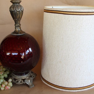 May include: A vintage table lamp with a dark red glass globe base and a white fabric lampshade with a brown trim. The lamp has a gold metal base with ornate details.