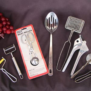 May include: A set of nine silver kitchen tools, including a meat baller, a slotted spoon, a garlic press, a pizza cutter, a peeler, and a strainer. The tools are arranged on a dark gray surface.