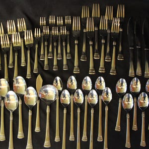 May include: A collection of silverware, including forks, spoons, and knives, arranged on a black surface. The silverware has a gold-colored handle and a silver-colored head. The knives have a black handle and a silver blade.
