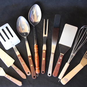May include: A collection of stainless steel and wooden kitchen utensils, including spatulas, spoons, a whisk, and a fork, arranged on a black surface. The utensils have wooden handles and are designed for cooking and food preparation.