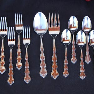 May include: A collection of silverware, including forks and spoons, with ornate handles. The silverware is arranged on a dark surface, reflecting the light. The handles have a brown and silver design.
