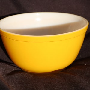 May include: A yellow glass mixing bowl with a white interior. The bowl is sitting on a dark brown surface.