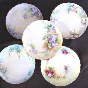 May include: Five antique porcelain plates with floral designs. Each plate features a different arrangement of flowers in shades of purple, pink, and blue, with gold trim. The plates are arranged on a dark background, showcasing their delicate artwork.