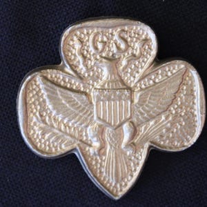 May include: A gold-colored Girl Scout pin featuring an eagle with outstretched wings holding a shield with the American flag. The eagle is surrounded by a wreath of leaves and the pin is shaped like a four-leaf clover.