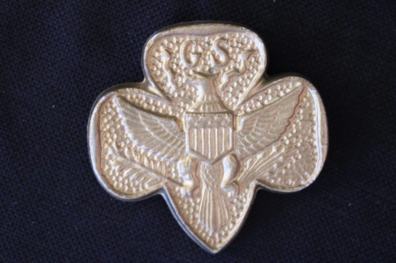 May include: A gold-colored Girl Scout pin featuring an eagle with outstretched wings holding a shield with the American flag. The eagle is surrounded by a wreath of leaves and the pin is shaped like a four-leaf clover.