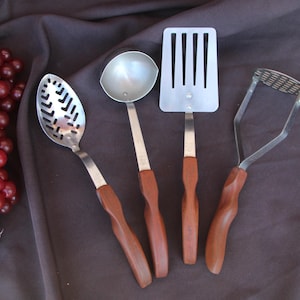 May include: A collection of four kitchen utensils: a slotted spoon, a ladle, a spatula, and a potato masher. Each tool has a stainless steel head and a wooden handle. The utensils are arranged on a dark brown fabric.