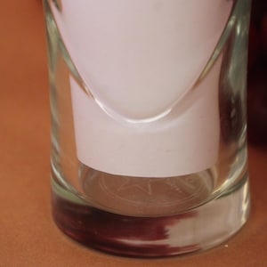 May include: A clear glass tumbler with a cylindrical shape and a thick base. The glass features a subtle, etched star design on the bottom interior. A white paper insert is placed inside the glass, and dark red grapes are visible to the right.