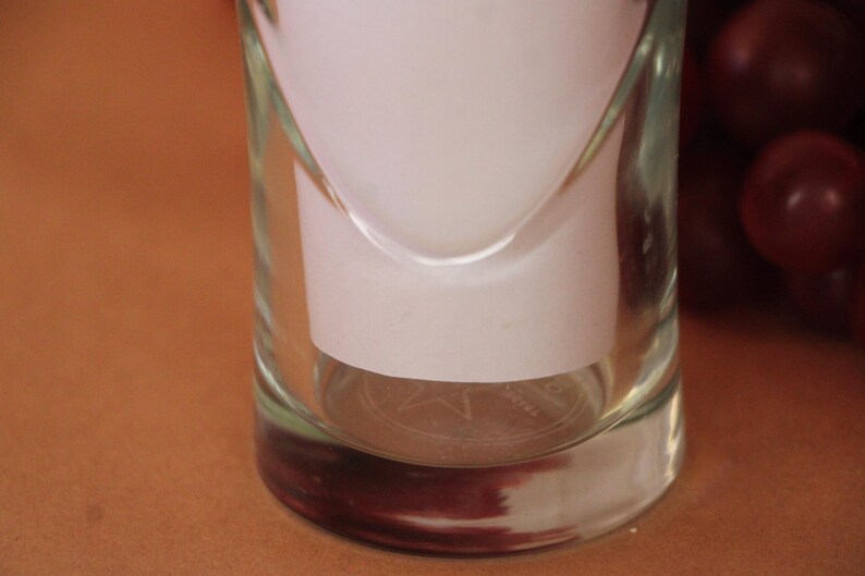 May include: A clear glass tumbler with a cylindrical shape and a thick base. The glass features a subtle, etched star design on the bottom interior. A white paper insert is placed inside the glass, and dark red grapes are visible to the right.