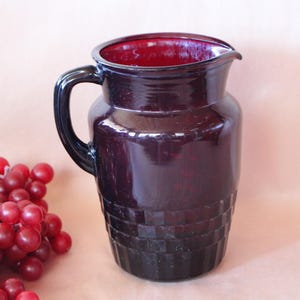 May include: A deep red glass pitcher with a smooth upper body and a textured, checkered pattern on the lower half. The pitcher features a sturdy handle and a small pouring spout. Beside the pitcher lies a cluster of artificial red grapes.