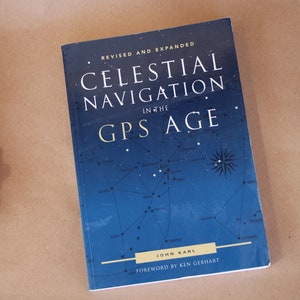 May include: A blue book with the title "Celestial Navigation in the GPS Age" by John Karl. The cover features a star chart with a compass rose.